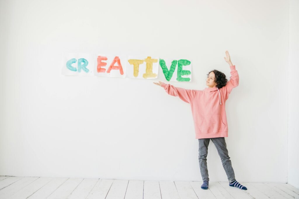 A child with curly hair in a pink hoodie points to colorful 'CREATIVE' text on a white wall.
